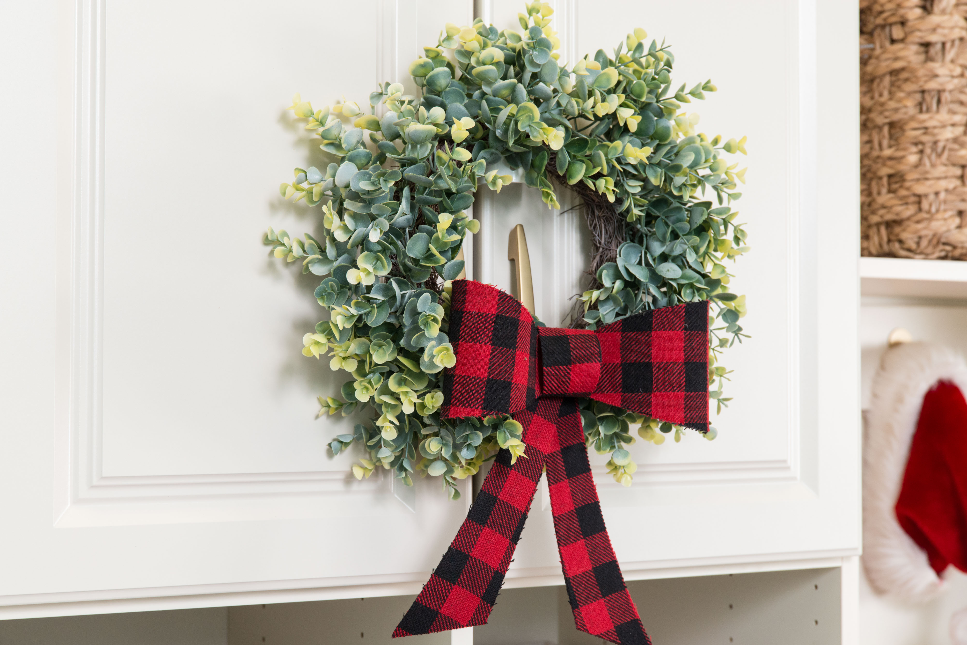 Wreath decor on cabinet with red and black buffalo checkered ribbon and Santa hat in background.